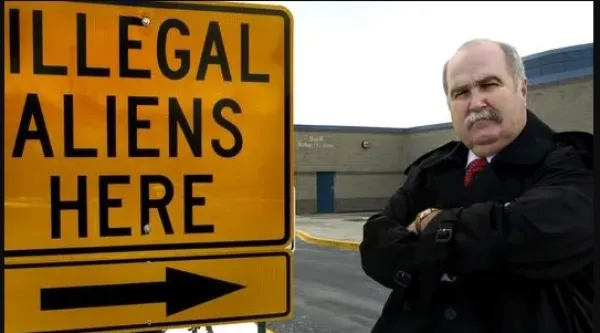 SIgn saying illegal aliens here and a stern bald older man with arms crossed and