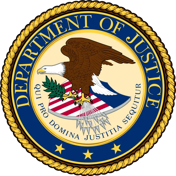 Round official seal with gold braided outside rim and blue around the circle that has words Department of Justice and an eagle facing left with wings held high on top of a shield with flag colors red, white and blue,