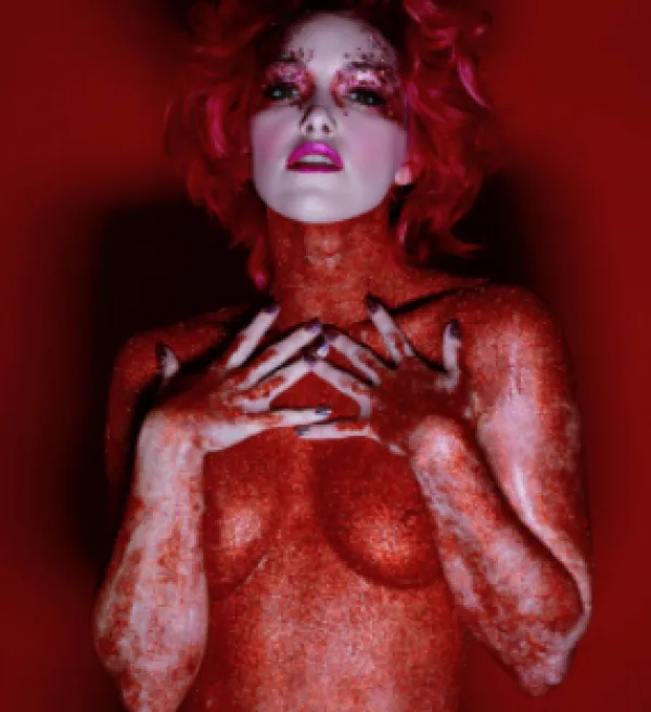 A woman looking like she's painted red, nude with her arms up across her breasts and hands with fingers spread near her throat. Her face is painted pure white and she has on a lot of makeup. Her hair is blonde, short and curly and the background is red.