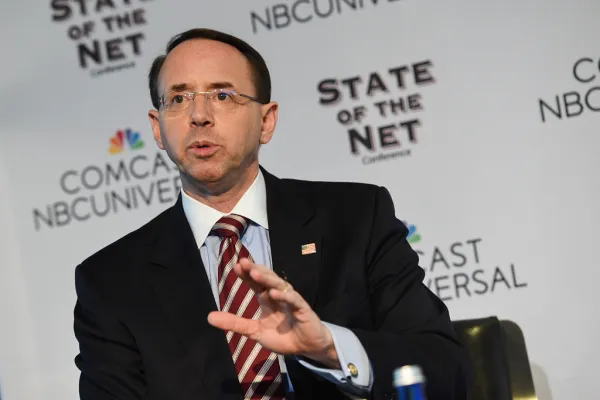 Balding white man with wire rimmed glasses wearing a suit speaking and make a hand gesture