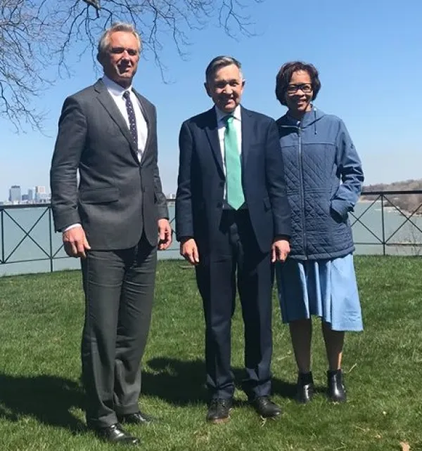Three people standing outside on the grass with body of water behind them, a tall white man in a suit with gray hair, and shorter white man in a suit and a black woman with glasses