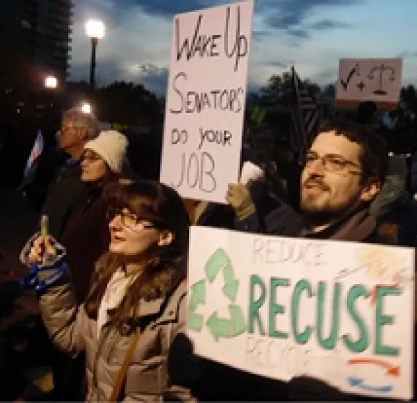 People standing outside in winter coats at dusk one white man with brown hair, facial hair and glasses with a sign that reads Recuse and a white woman next to him